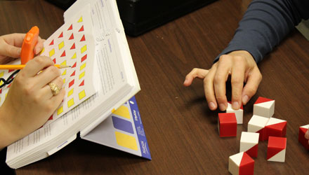 A person arranging red and white blocks with another person looks at a manual